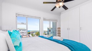 Un dormitorio con cama, ventilador de techo, ventanas amplias y vista a edificios.