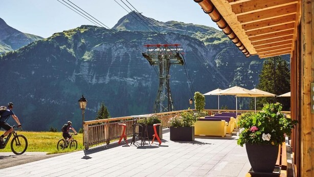 Blick auf Berge mit einer Sesselbahn, Sitzgelegenheiten im Freien und einem Holzhaus.