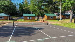 A tennis court with a net, surrounded by green spaces and wooden cabins.