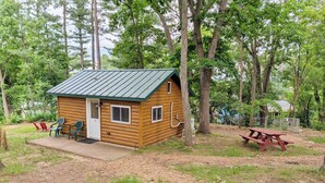 A small wooden cabin with a metal roof, a door, and two windows. There is a picnic table and two chairs outside.