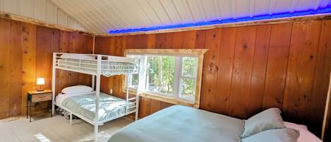 Un dormitorio tipo cabaña con una litera, una cama individual, un ventanal con vistas a la vegetación y un techo iluminado en azul.