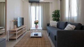 Ett modernt vardagsrum med en grå soffa, ett vitt soffbord, en tv på ett vitt tv-bord och ett stort fönster med gardiner.