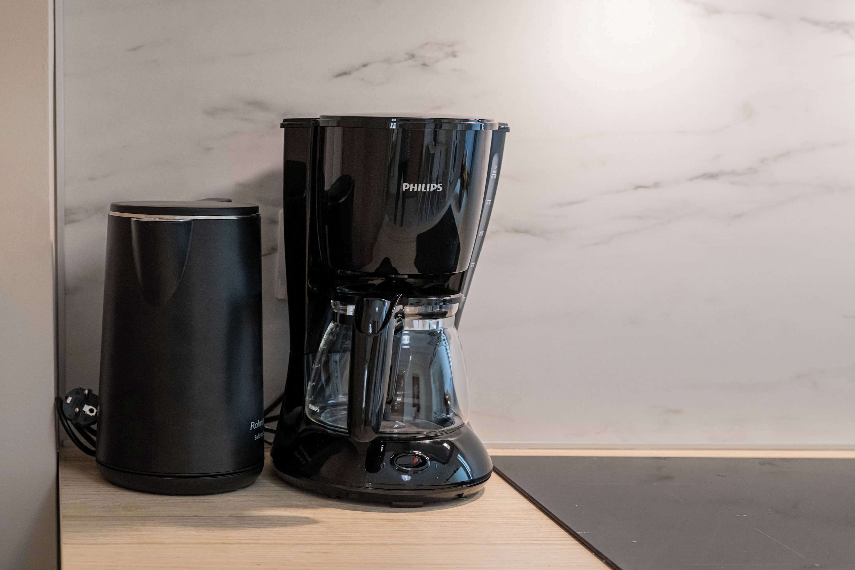 A black coffee maker and a black thermos on a wooden surface against a marble wall.