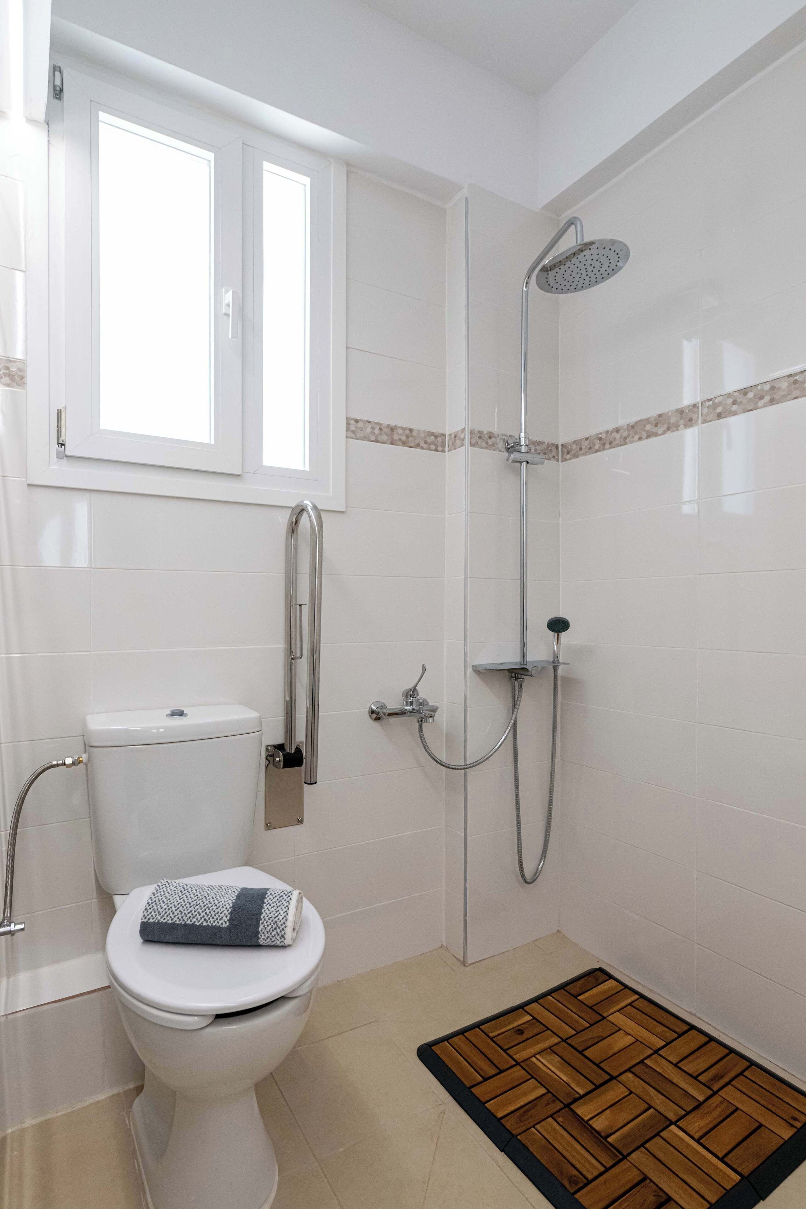 A bathroom with a toilet, a shower with a handheld showerhead, and a window above the toilet.