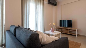 Un salon moderne avec un canapé gris, un téléviseur à écran plat posé sur un meuble en bois, une petite table blanche ornée d’un vase de fleurs et une grande fenêtre avec des rideaux.