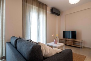 Un moderno salón con un sofá gris, un televisor de pantalla plana sobre un mueble de madera, una pequeña mesa blanca con un florero y un amplio ventanal con cortinas.