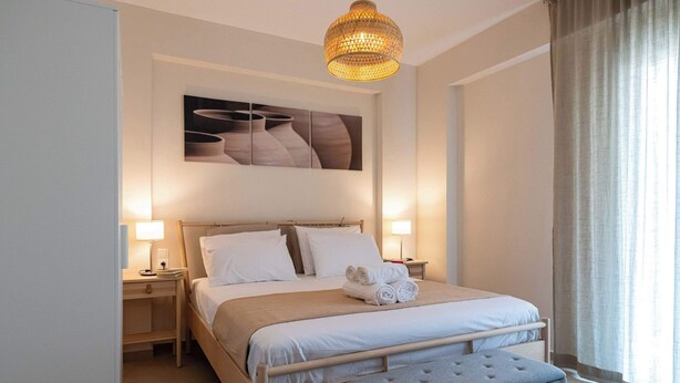 Un lit bien fait, avec du linge de lit blanc, un banc recouvert d’un tissu gris et une œuvre d’art accrochée au mur au-dessus.