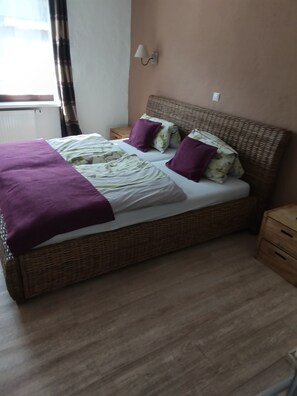 Un lit avec une couverture violette, une couette blanche et deux oreillers violets. Il y a une table de chevet avec une lampe et un tiroir en bois.