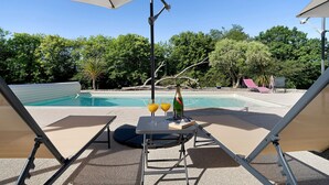 Uma área na beira da piscina com espreguiçadeiras, uma mesa com bebidas e uma piscina ao fundo.