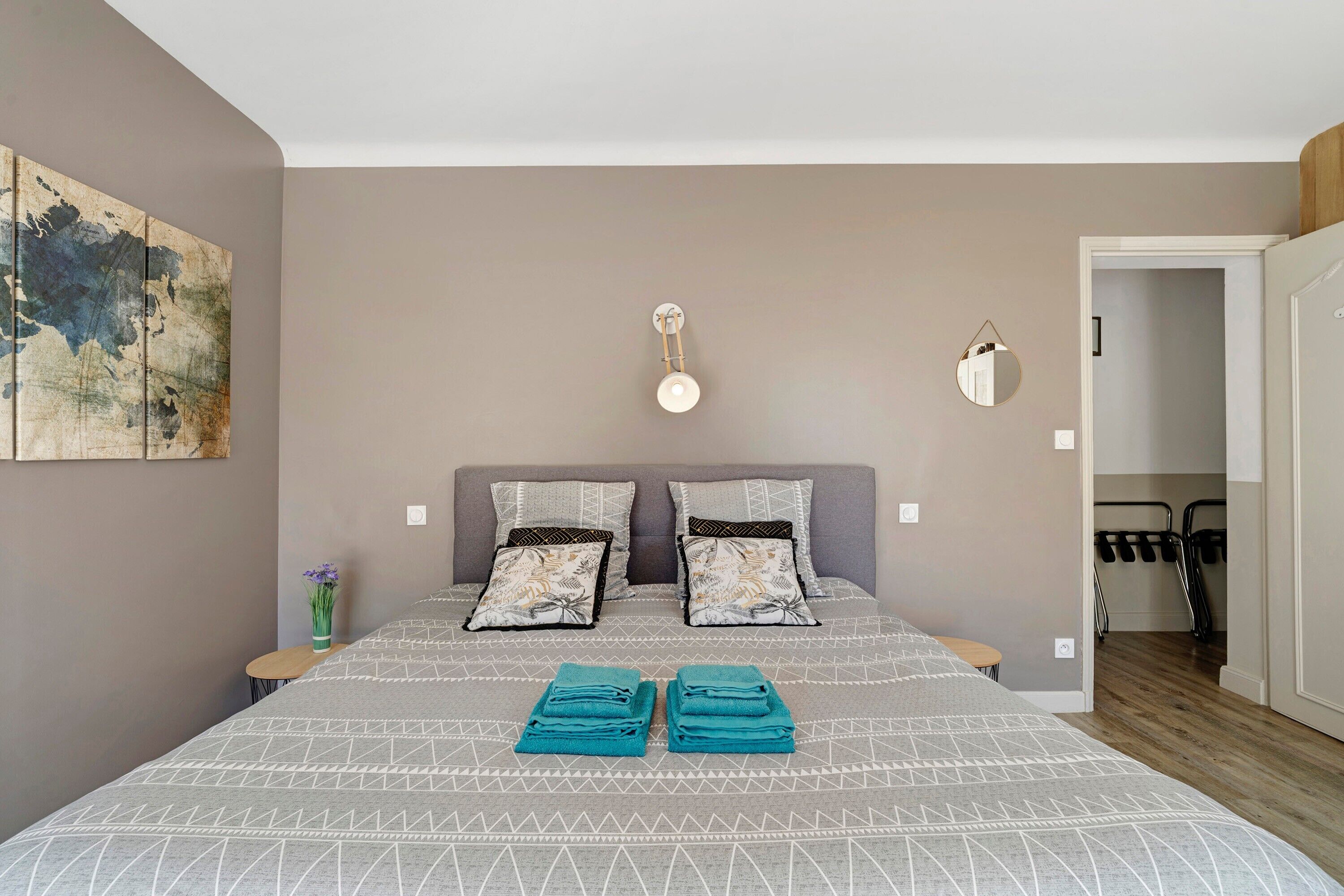 A bedroom with a bed, pillows, a wall clock, a mirror, and a vase with flowers.