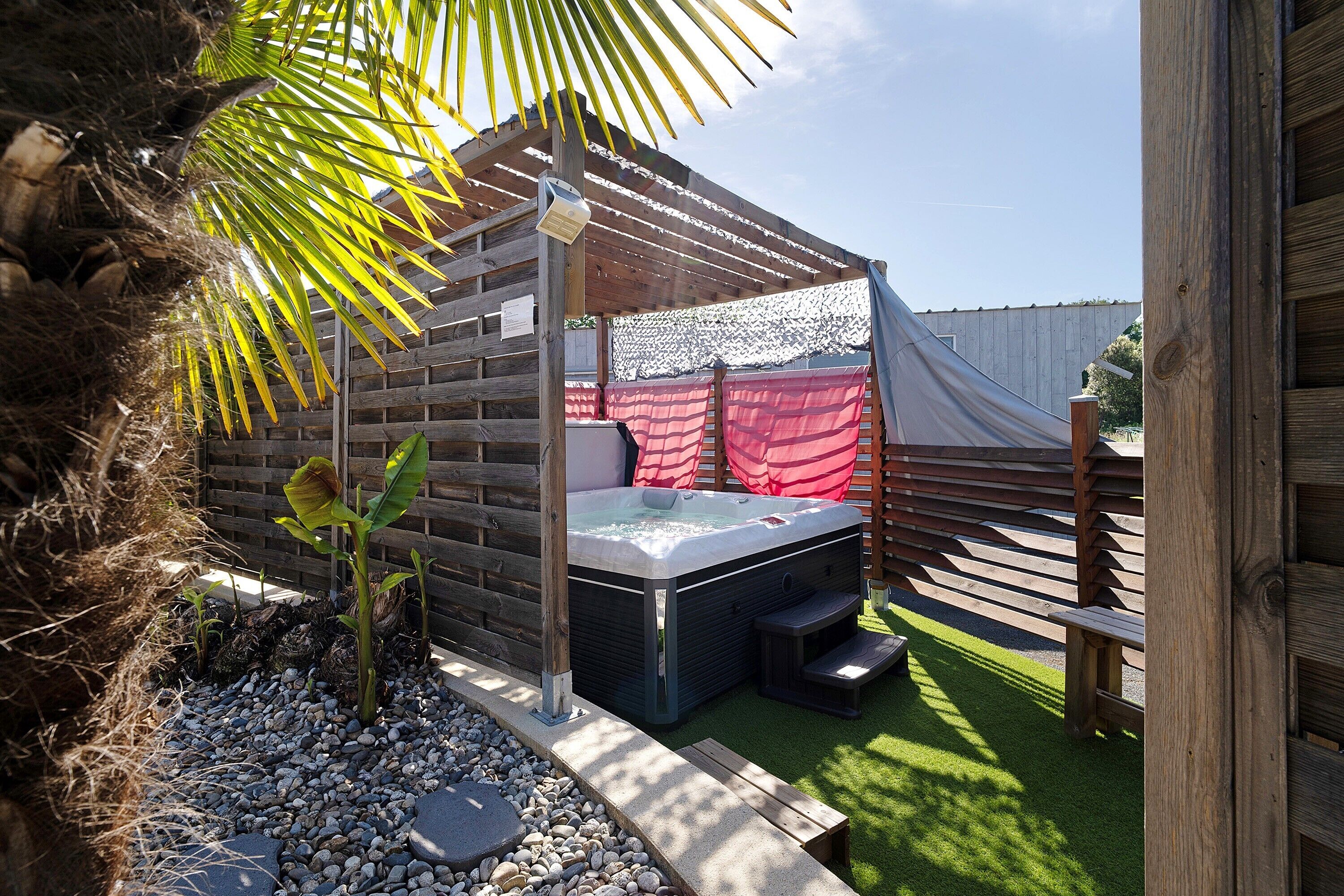 A hot tub in a wooden structure with a wooden fence, surrounded by pebbles and a small plant.