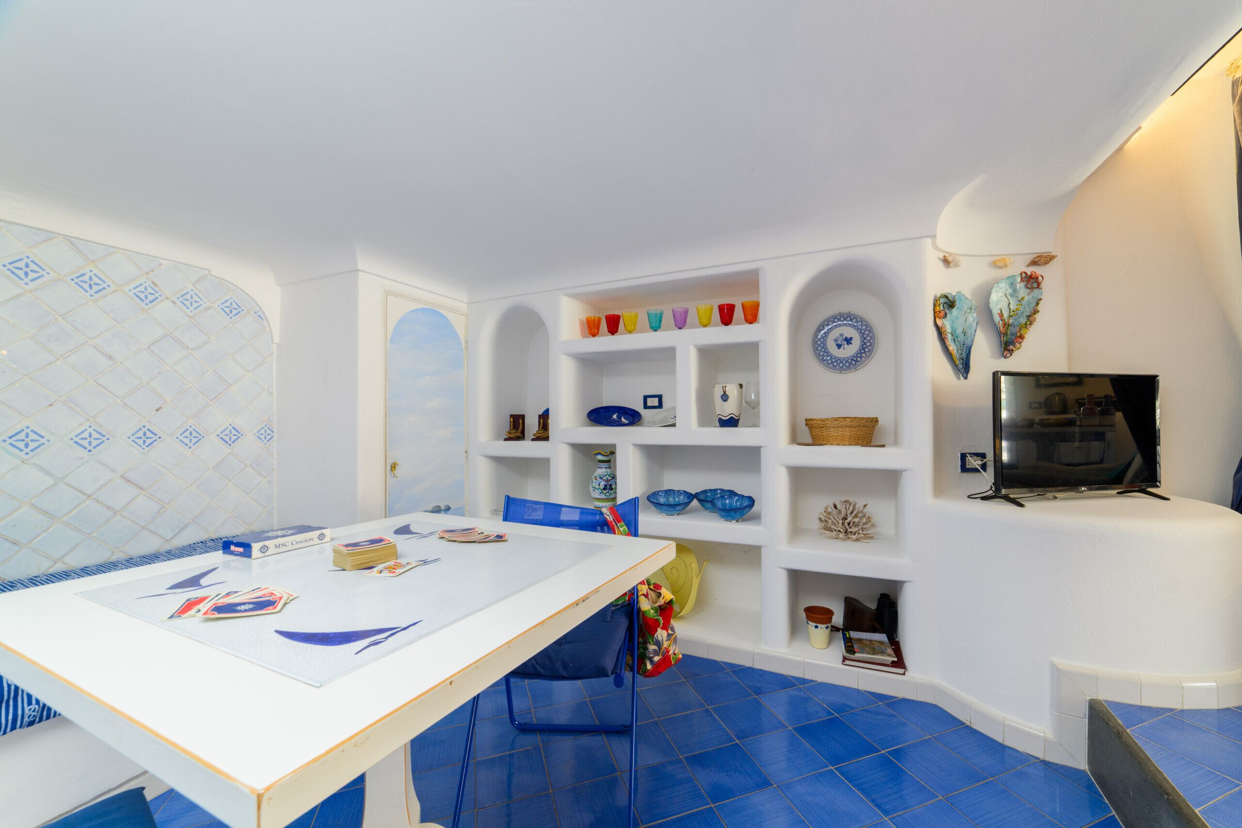 Ein Zimmer mit einem weißen Tisch, blauen Stühlen und einem Fernseher. Außerdem sind Regale mit Dekoration und eine Wand mit Muster zu sehen.