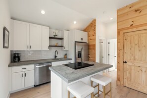 A modern kitchen with white cabinets, a stainless steel refrigerator, a built-in oven, and a grey countertop island with stools. - The Keowee Hideaway• Lakefront • 25 min to Clemson (Seneca)