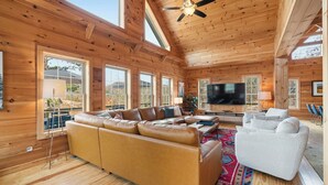 A spacious living room with a large leather sofa, a white armchair, and a flat-screen TV. The room features wooden walls and a high ceiling with a fan. - The Keowee Hideaway• Lakefront • 25 min to Clemson (Seneca)