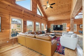 A spacious living room with a large leather sofa, a white armchair, and a flat-screen TV. The room features wooden walls and a high ceiling with a fan. - The Keowee Hideaway• Lakefront • 25 min to Clemson (Seneca)