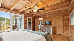 A wooden cabin bedroom with a large bed, a ceiling fan, a TV, and a sliding glass door leading to an outdoor area. - The Keowee Hideaway• Lakefront • 25 min to Clemson (Seneca)