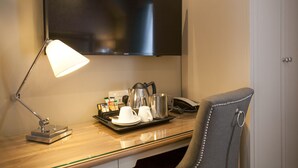 Habitación de hotel con escritorio, televisor de pantalla plana montado en la pared, lámpara de escritorio, silla y bandeja con tazas de café y cafetera.