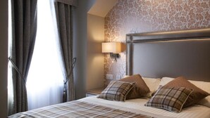 Habitación de hotel con una cama grande, un cabecero con lámpara, una ventana con cortinas y una pared con diseño floral.