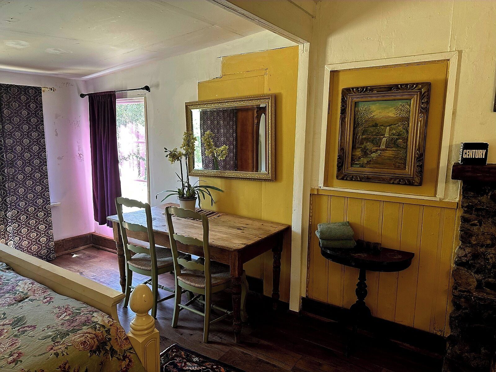 A room with a bed, a wooden table, two chairs, a potted plant, a mirror, and a framed painting on the wall.