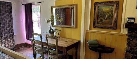 A room with a bed, a wooden table, two chairs, a potted plant, a mirror, and a framed painting on the wall.