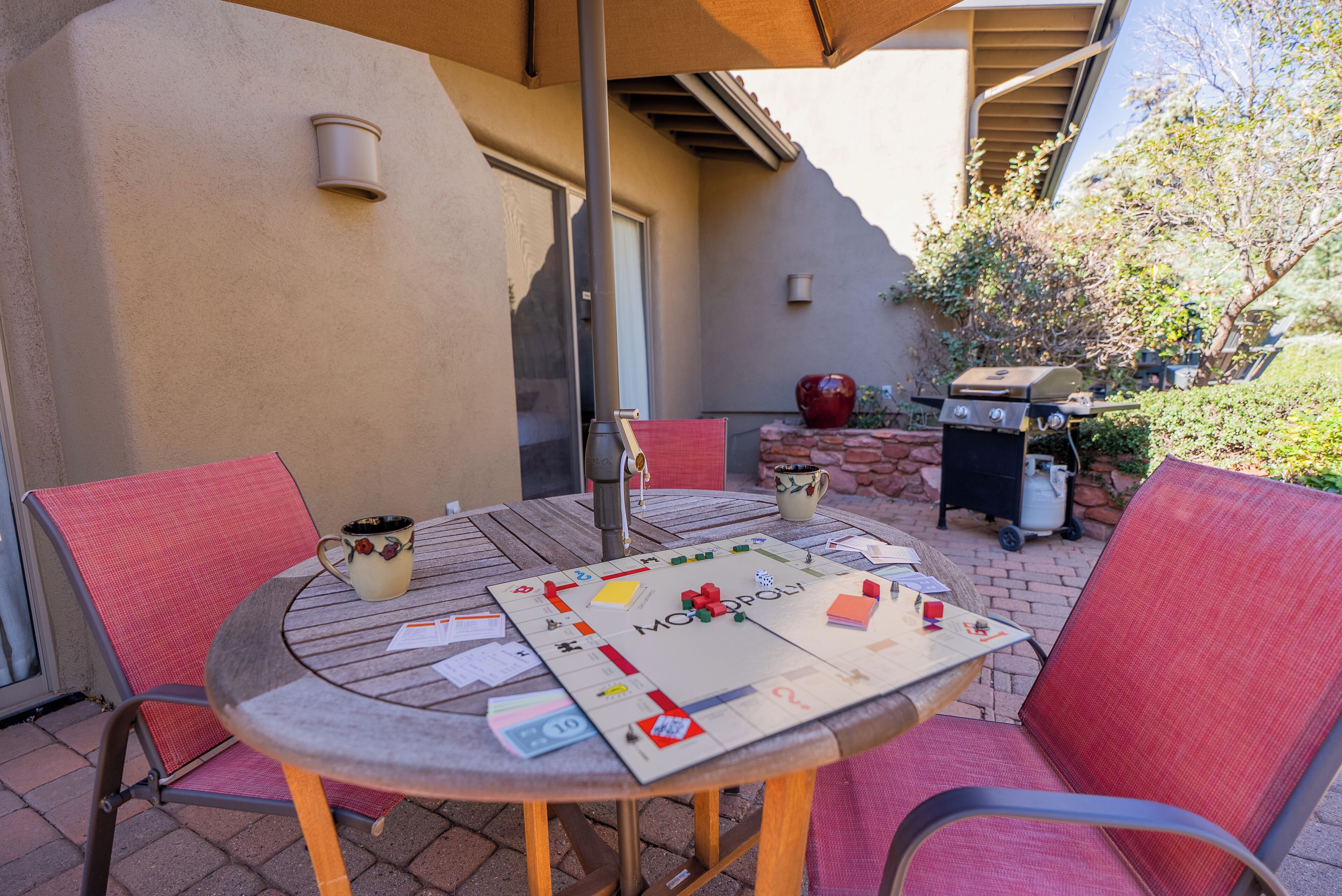 A round table with a Monopoly board game, two chairs, a coffee mug, and a grill in the background.