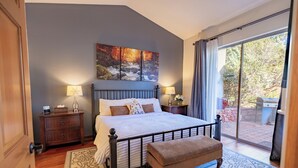 Un dormitorio con una cama grande, dos mesitas de noche con lámparas, un banco, un tapete estampado y un cuadro con un paisaje.
