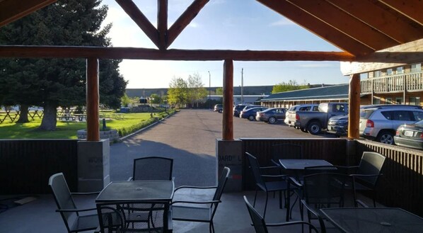Un espace extérieur aménagé en terrasse, avec des tables et des chaises, une allée couverte et une vue sur un parking rempli de voitures.