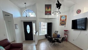 Ein geräumiges Wohnzimmer mit einem roten Sofa, einem Essbereich mit Tisch und Stühlen, einem an der Wand montierten Fernseher und einem großen Fenster mit Jalousien.