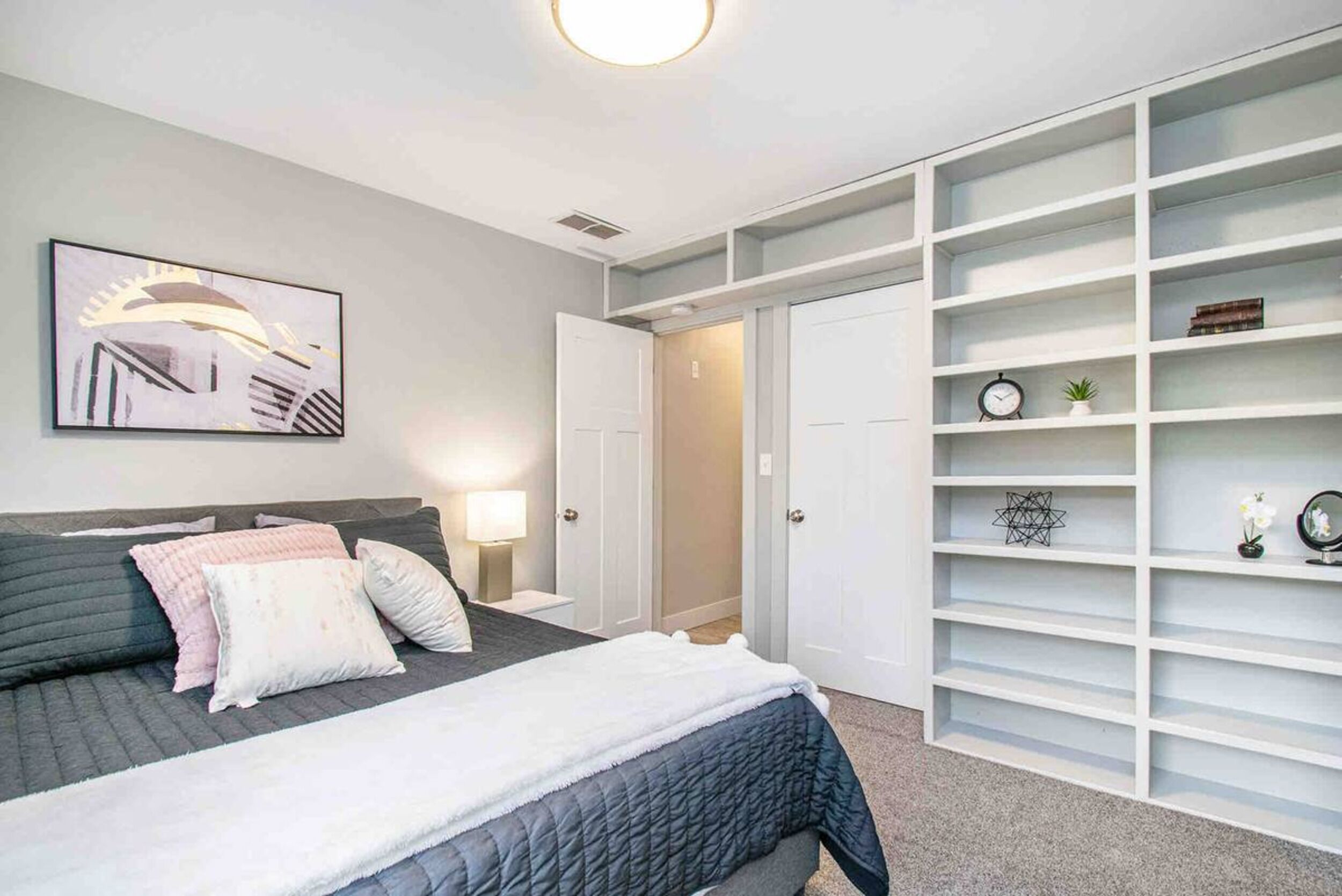 A bedroom with a bed, bedside tables, a nightstand, a wall-mounted artwork, and a built-in shelving unit.