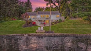 A two-story house with a balcony, surrounded by a well-maintained lawn and a body of water in the foreground.