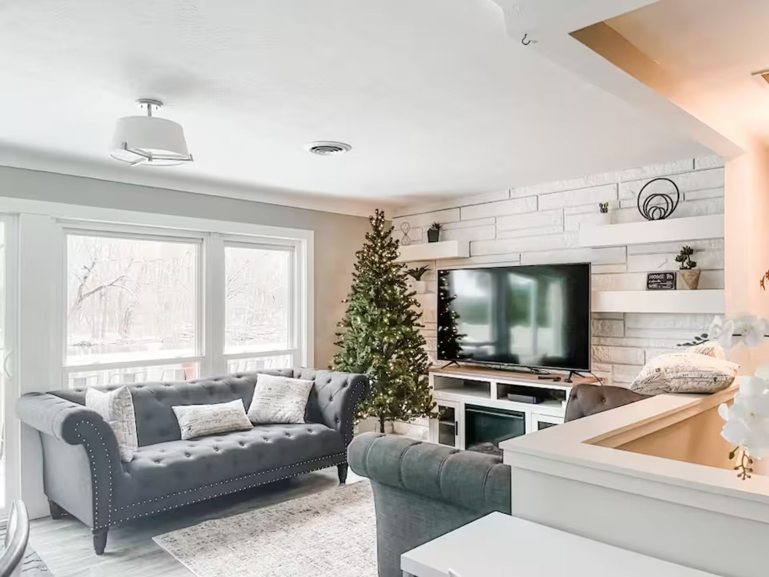 Una sala de estar moderna con un sofá gris, un sillón verde pequeño, una mesa de centro blanca, un televisor de pantalla plana y un árbol de Navidad.