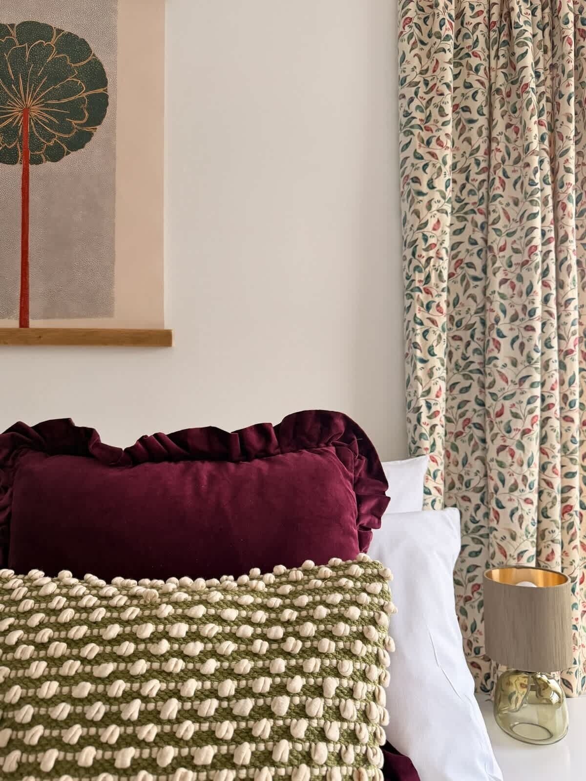 A neatly made bed with a green and white patterned blanket, a dark red pillow, and a white pillow. There is a framed artwork on the wall and a lamp on the bedside table.