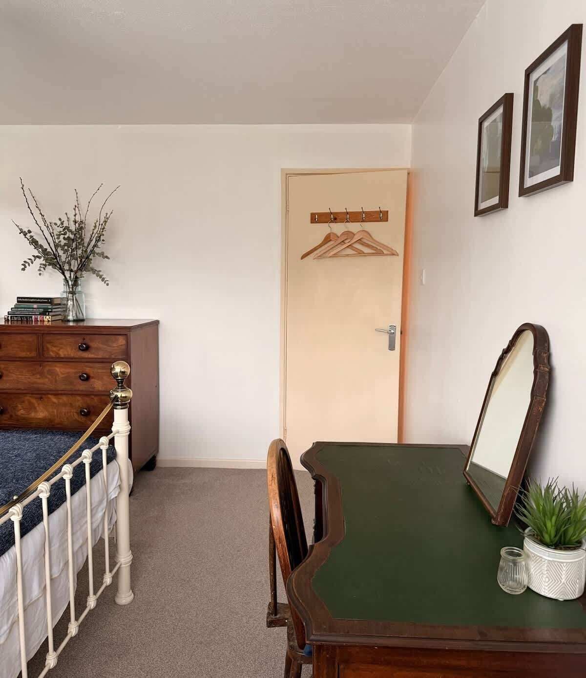 A bedroom with a bed, dresser, chair, table, mirror, and a door with a coat rack.