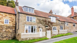 Une maison en pierre avec une clôture en bois peinte en blanc, une baie vitrée en forme d’arc et un toit recouvert de tuiles rouges.