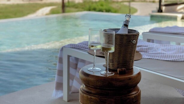 A wooden table with a bottle and two glasses, overlooking a pool with a view of the beach and palm trees.