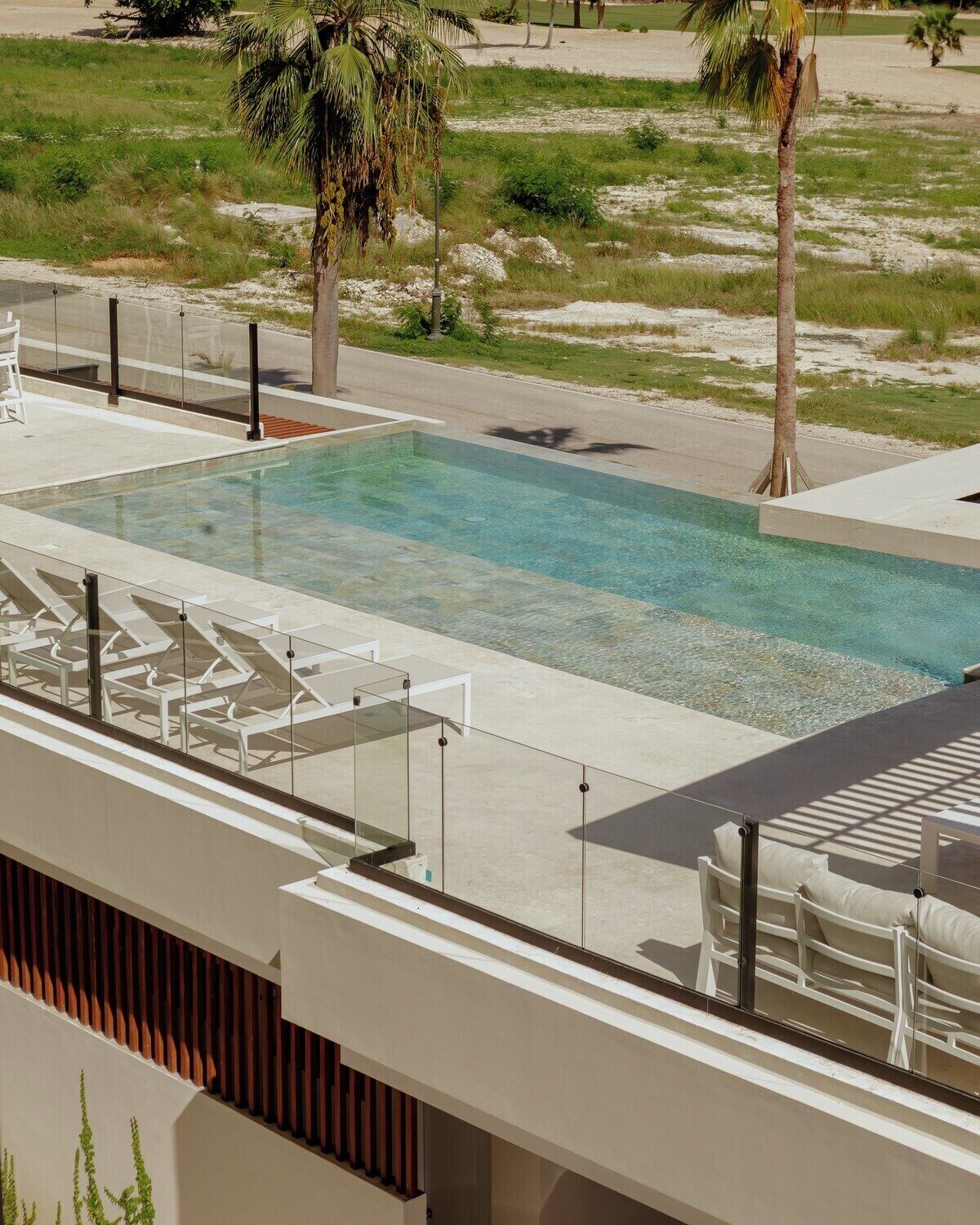 A rectangular swimming pool with clear water, surrounded by a concrete deck. There are lounge chairs and a glass railing along the pool's edge. Palm trees are visible in the background.