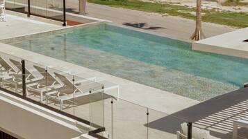 A rectangular swimming pool with clear water, surrounded by a concrete deck. There are lounge chairs and a glass railing along the pool's edge. Palm trees are visible in the background.