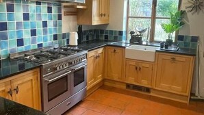 A kitchen with a gas stove, oven, wooden cabinets, and a window with a view of greenery. - Spacious Village House near Oxford with Woodburner (Horspath)