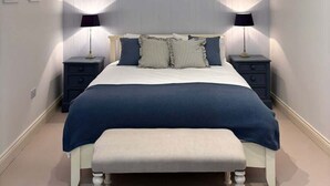 A bedroom with a large bed, two bedside tables with lamps, a bench at the foot of the bed, and a sloped ceiling with recessed lighting.