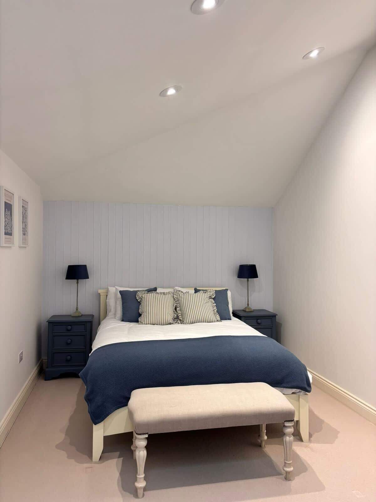 Un dormitorio con una cama, dos mesitas de noche, un banco e iluminación empotrada en el techo.