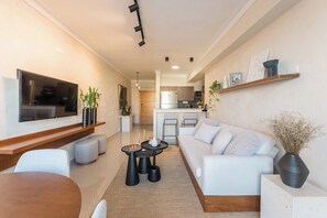 Uma sala de estar moderna com um sofá, mesa de centro e televisão. Cozinha visível ao fundo, com geladeira e micro-ondas.