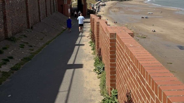 A coastal walkway with a glass structure, a brick wall, and a beach in the background.