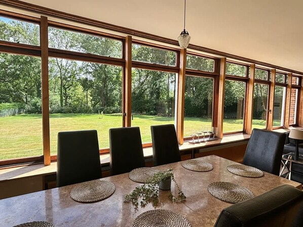 A dining area with a large table, four chairs, and a view of a grassy yard through large windows.
