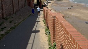 Ein Küstenweg mit einer Glasgalerie, einer Backsteinmauer und einem Strand im Hintergrund.