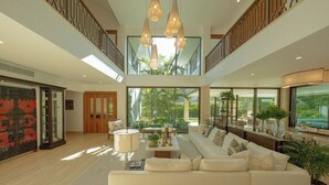 Ein geräumiges Wohnzimmer mit einer großen Couch, einem Couchtisch und einer Chandelüster. Eine Treppe führt zu einer oberen Ebene, und große Fenster sorgen für natürliches Licht.