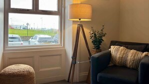 En hyggelig stue med en mørkeblå sofa, en rund vævet skammel, en gulvlampe og et vindue med udsigt til en parkeringsplads.