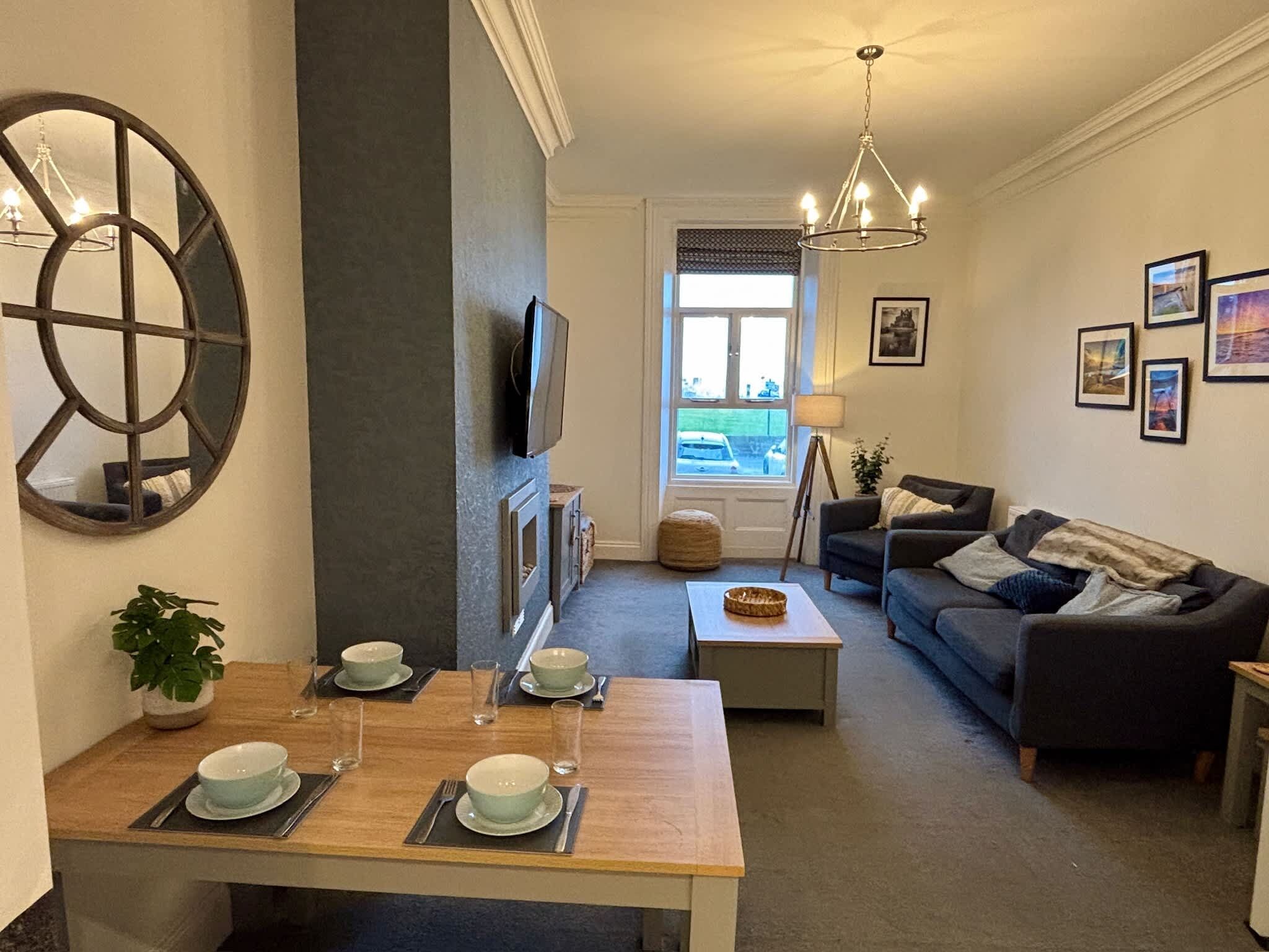 Una sala de estar moderna con un sofá, una mesa de comedor para dos personas, un televisor montado en la pared y un ventanal con vistas al exterior.