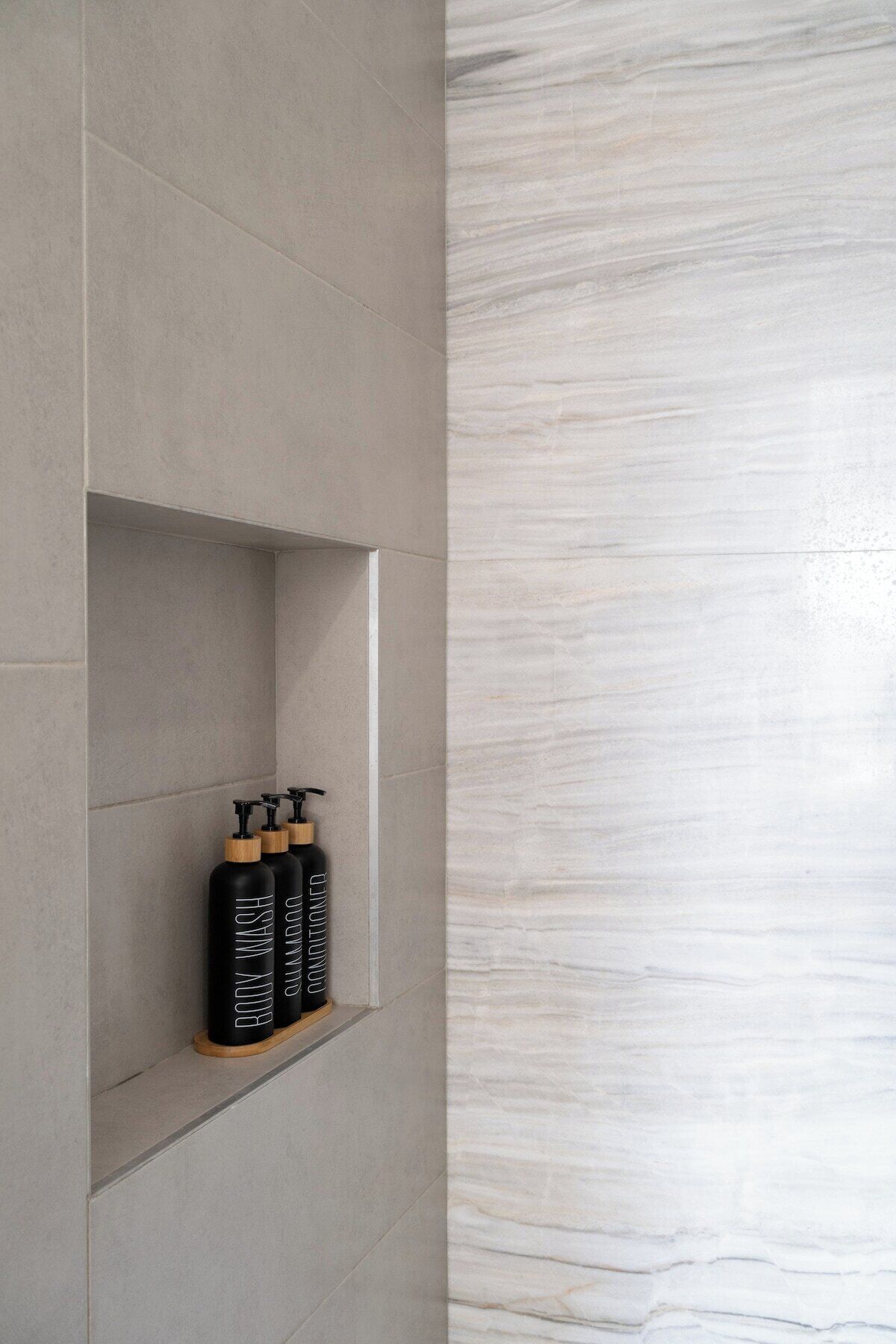 A bathroom with a grey tiled wall and a marble-like textured wall. There is a shelf with three black bottles labeled "BODY WASH" and "SHAMPOO" on the grey wall.