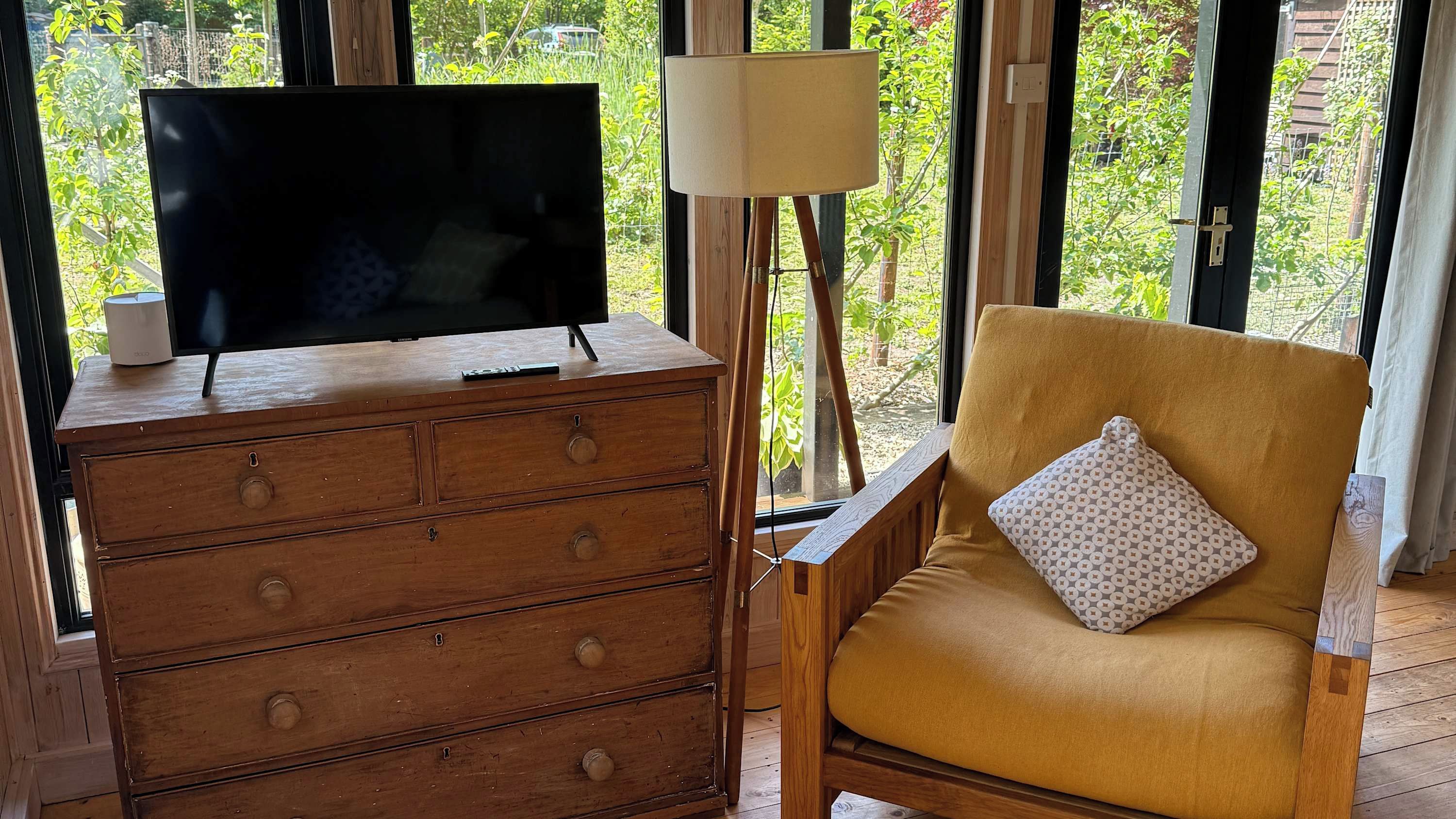 Eine Holzkommode mit einem Flachbildfernseher, einer Lampe und einem gemusterten Kissen auf einem Stuhl.