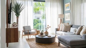 Een moderne woonkamer met een grijze bank, een ronde salontafel en een televisie op een houten dressoir. Er is een groot raam met dunne gordijnen, een kamerplant en een ingelijst schilderij aan de muur.
